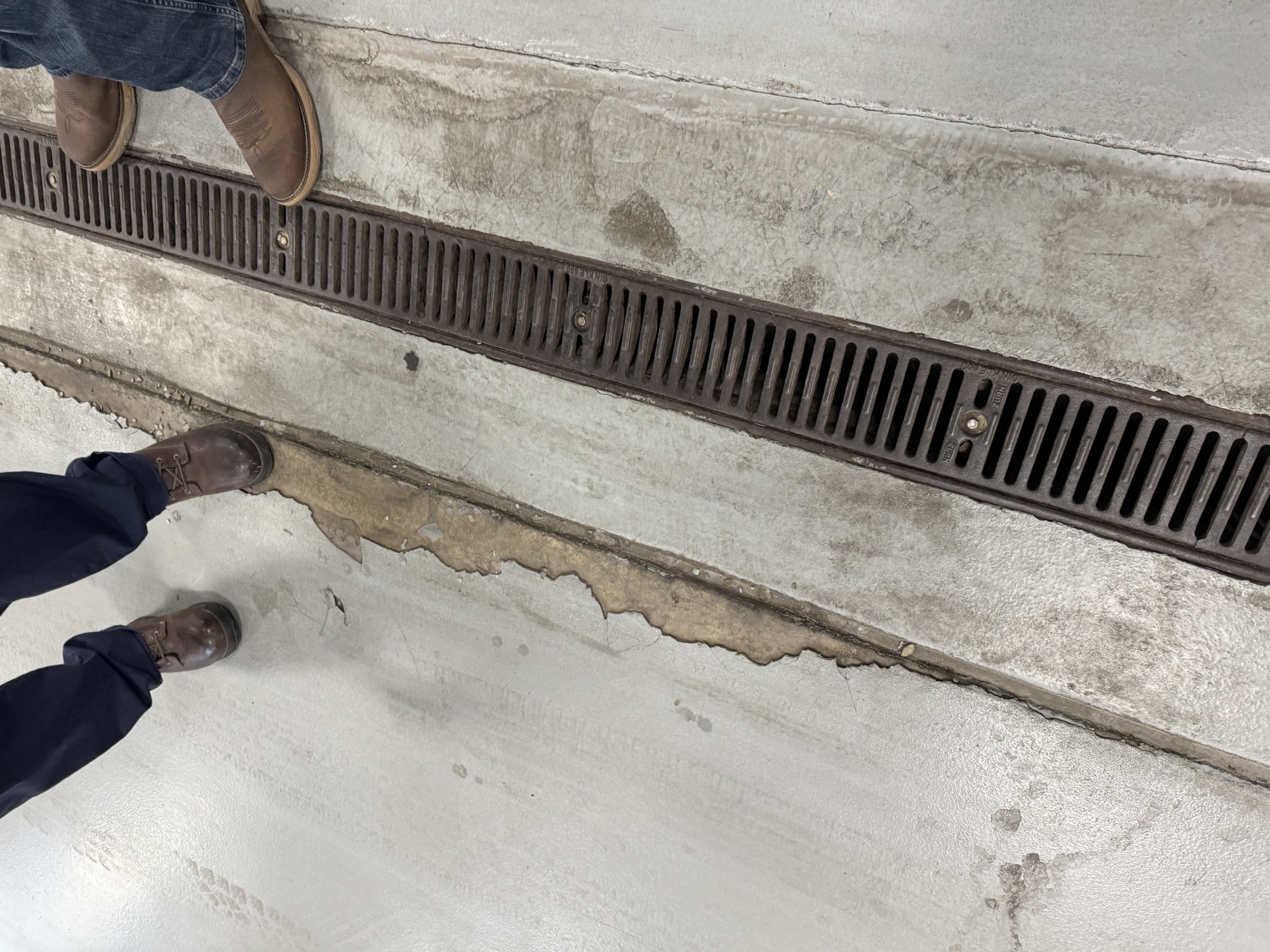 Damage survey: the old coating peeling and delaminating along the trench drain edge, with oil staining soaked into the exposed concrete.